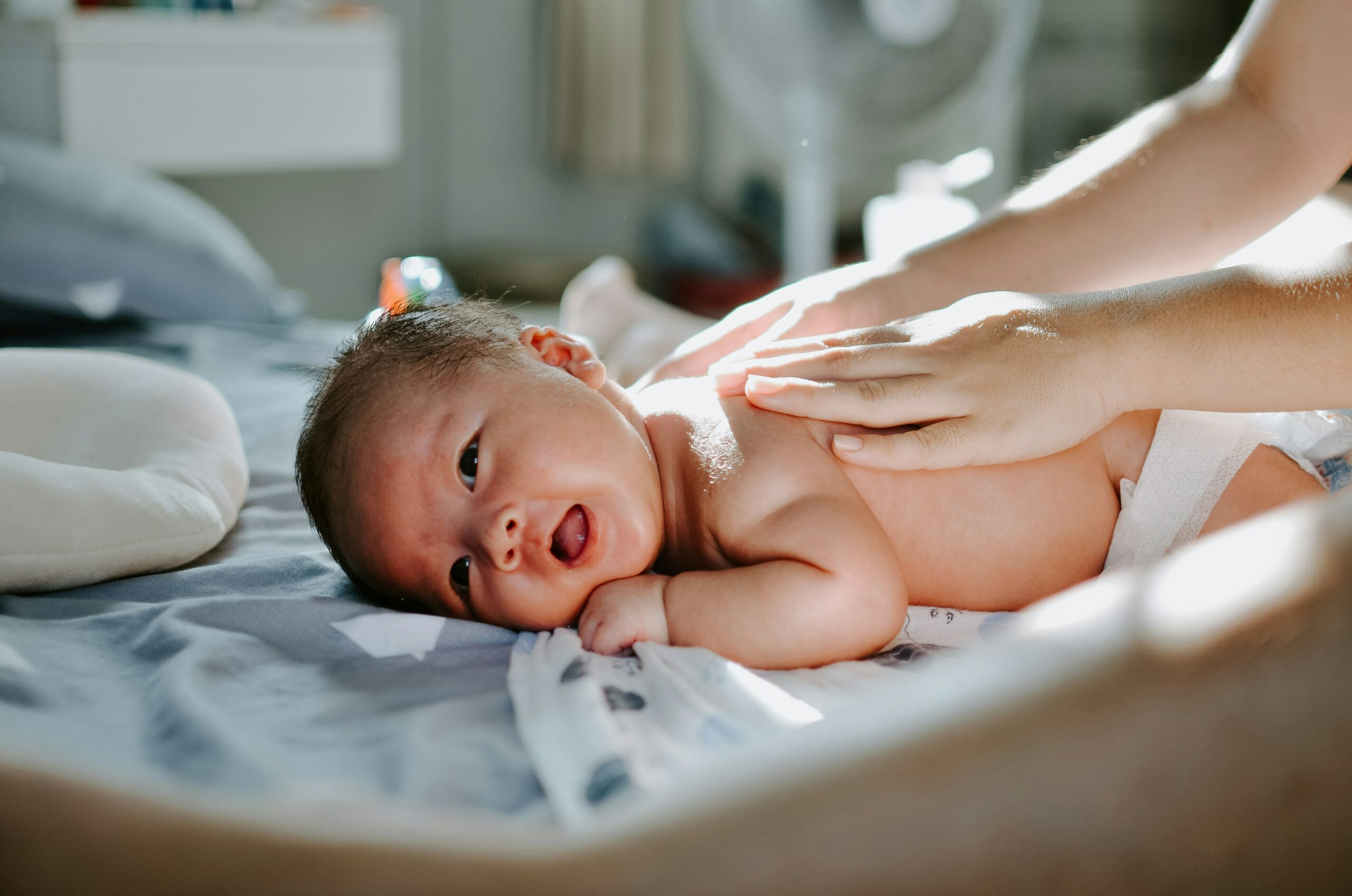 photo d'un bébé éveillé et souriant, avec deux mains posées sur lui, comme pour lui prodiguer un massage.